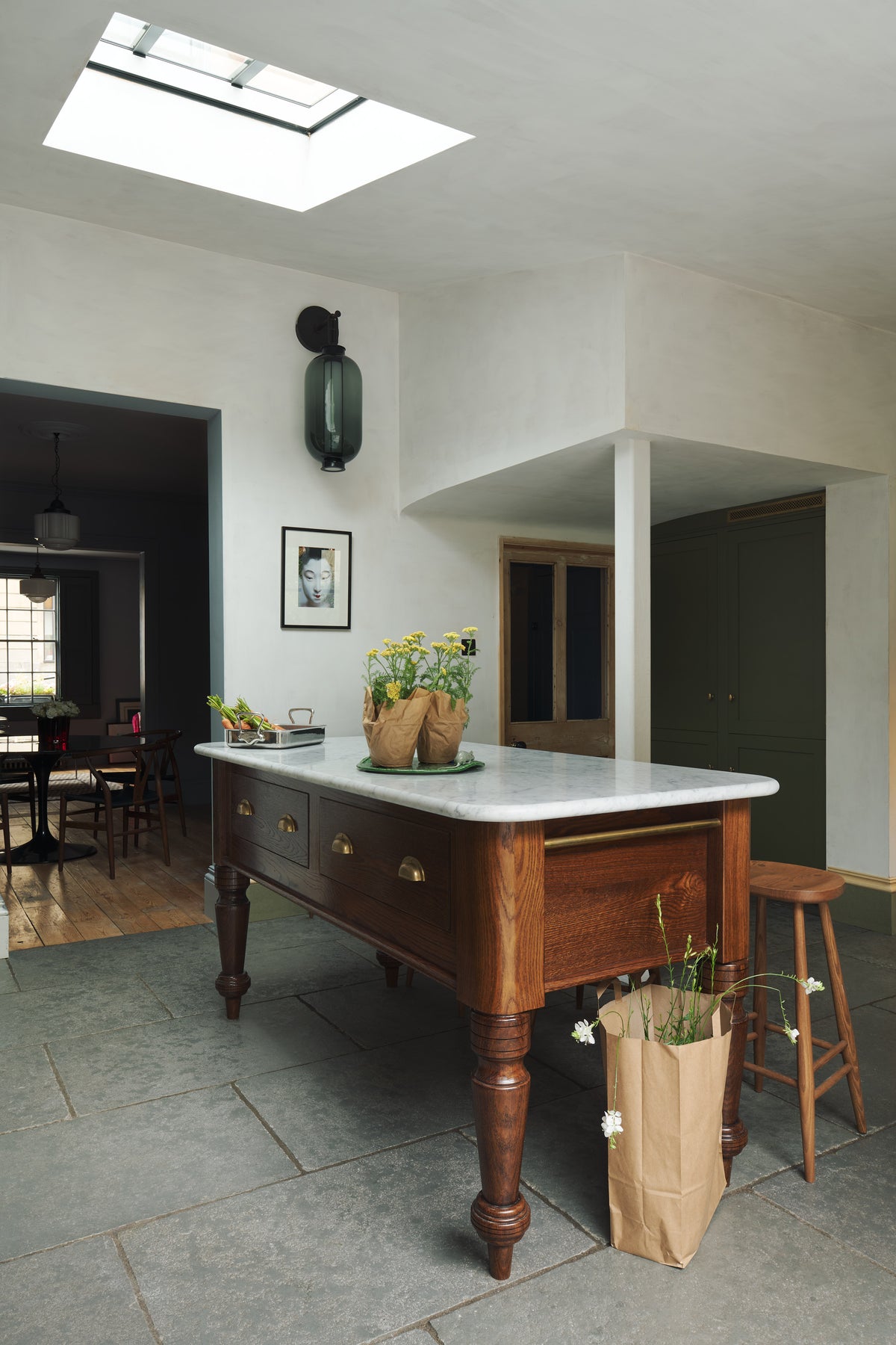 An oblique view of a quaint and modern Wilkes green Shaker kitchen features a Dairy table with a breakfast nook. Adorned with potted flowers, sky light, bum stools, and a signature glass chinese lantern inspired light it embodies a playful industrial and practical design.