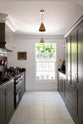 Looking through a simple archway is a murky-green galley shaker kitchen, featuring a cooker run with a black granite worktop and splashback, an aged brass hanging hook, a shaker canopy, a sink run, and pantry cupboards. Beautifully curated with two copper pendant lights, a vintage gold-framed painting, artisan ceramics, fresh produce, houseplants, and a wide open window, it creates a chic, understated, and practical galley kitchen.