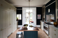 A simple and elegant mushroom-brown Shaker kitchen features a cooker run with freestanding cupboards, a Carrara marble worktop, a large kitchen island with a single Belfast sink, a small section of copper worktop, and a bank of fitted cupboards housing a fridge-freezer and storage pantries. Adorned with artisan ceramics, a playful framed illustration, gem lettuce, bright red rhubarb, luscious houseplants, and a deVOL Heirloom gaselier, the space feels loving and functional.