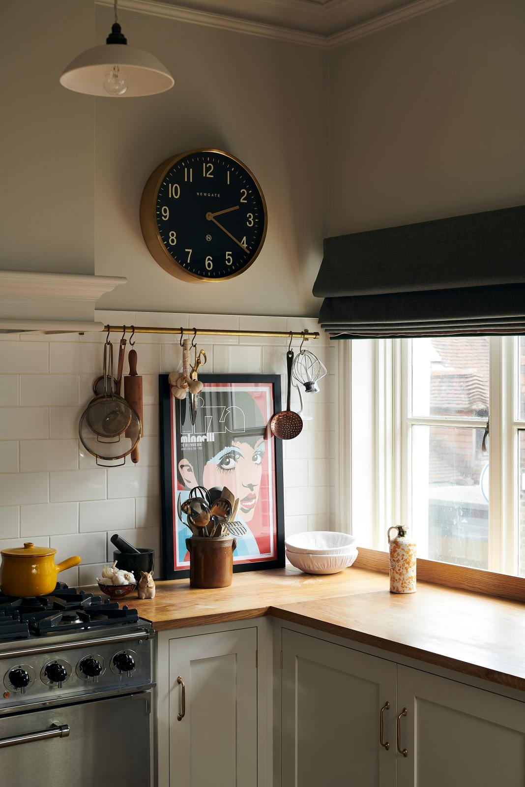 A corner of a vibrant Shaker kitchen features an L-shaped sink and cooker run with a solid oak worktop, lovingly decorated with a gold-rimmed clock, an aged brass hanging rail, a chattered skullcap porcelain light, beautiful ceramics, and a bold pop-art poster, creating a characterful and inviting space.
