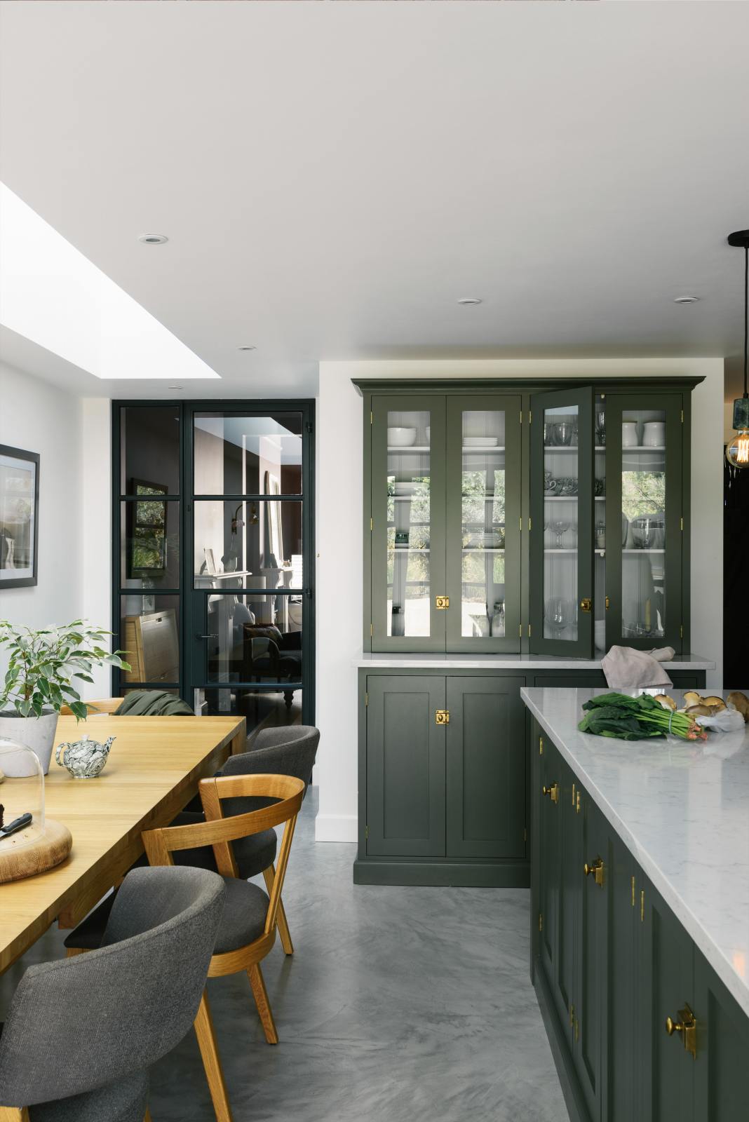 A bright and sophisticated Bakehouse Green shaker kitchen, styled with Scandinavian furniture and a soothing green palette. The kitchen features a central island with a double Belfast sink and a glazed larder with Silestone worktop. There’s a beautiful skylight above the solid oak dining table.