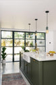 An angled view of a bright, sophisticated Bakehouse Green shaker kitchen, styled with Scandinavian furniture and a calming green palette. The kitchen showcases a central island with a double Belfast sink, Silestone worktop, and aged brass Ionian tap. Natural light floods the kitchen and dining room through the glass French doors.