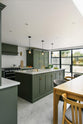 An angled view of a bright and sophisticated Bakehouse Green shaker kitchen, styled with Scandinavian furniture and a soothing green palette. The kitchen features a cooker run, a central island with a double Belfast sink and Silestone worktop, and full-height cupboards. Natural light floods the kitchen and dining room through the glass French doors.