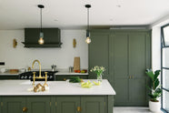 A bright and sophisticated Bakehouse Green shaker kitchen, styled with Scandinavian furniture and a soothing green palette. The kitchen features a cooker run, a central island with a double Belfast sink and Silestone worktop, and full-height cupboards. Natural light floods the kitchen and dining room through the glass French doors.