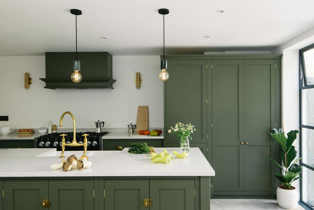 A bright and sophisticated Bakehouse Green shaker kitchen, styled with Scandinavian furniture and a soothing green palette. The kitchen features a cooker run, a central island with a double Belfast sink and Silestone worktop, and full-height cupboards. Natural light floods the kitchen and dining room through the glass French doors.
