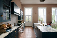The room, a grand Georgian space from the 18th century, features charming pink walls. It showcases a modern Clerkenwell Blue shaker kitchen equipped with glazed wall cupboards, a shaker-style canopy, a cooker run, and a spacious kitchen island with a sink and an Arabescato marble worktop. The kitchen offers views of the balcony and garden through its original wooden floors and doors, and the renovated space complements the esteemed property beautifully.