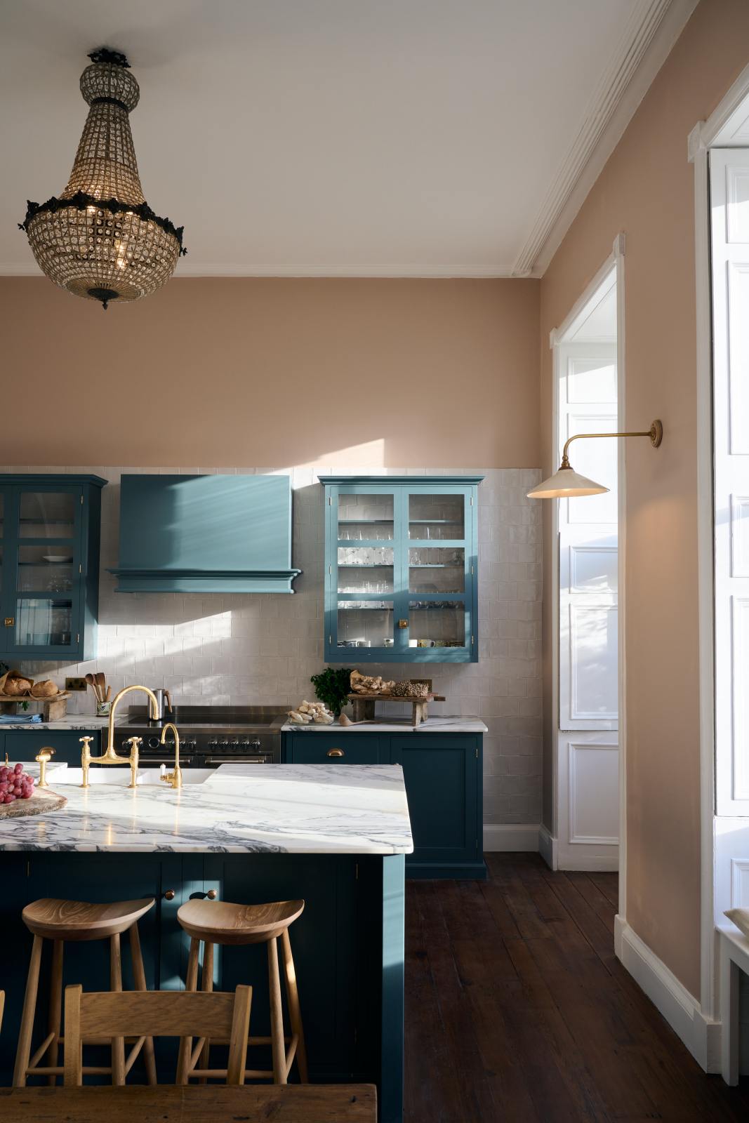 The room, a grand Georgian space from the 18th century, features charming pink walls. It showcases a modern Clerkenwell Blue shaker kitchen equipped with glazed wall cupboards, a shaker-style canopy, a cooker run, and a spacious kitchen island with a sink and an Arabescato marble worktop. Above is a beautiful vintage chandelier that enhances the renovated kitchen space and complements the esteemed property exceptionally well.