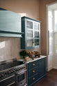 A closer look at a Clerkenwell Blue shaker kitchen’s cooker run equipped with Arabescato marble worktop, glazed wall cupboards, a shaker-style canopy and white porcelain splashback. Assorted fine drinking glasses and ceramic mugs are inside the glazed cupboard. On the worktop, there are herbs, bread, and fruits.