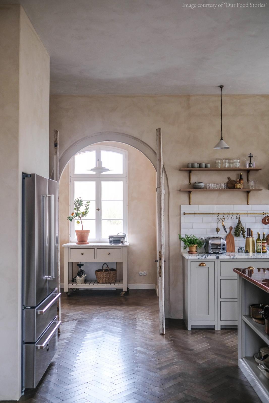 A grand yet grounded shaker kitchen in muted mushroom off-white, alive with scattered pots, pans, fruits, and vegetables. The L-shaped cooker and sink run features an Arabescato marble countertop, double-fluted sink, and aged brass Ionian tap. Crackle metro tiles and a long-aged brass rail form the splashback. A large copper-topped island and cosy breakfast nook complete the space.