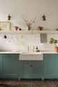 A close-up view of a Trinity Blue shaker kitchen sink features a Carrara marble countertop and a single, fluted Belfast sink with aged brass Mayan taps. Above the sink is a long shelf with brass rails for hanging cups, cleaning tools, and towels.