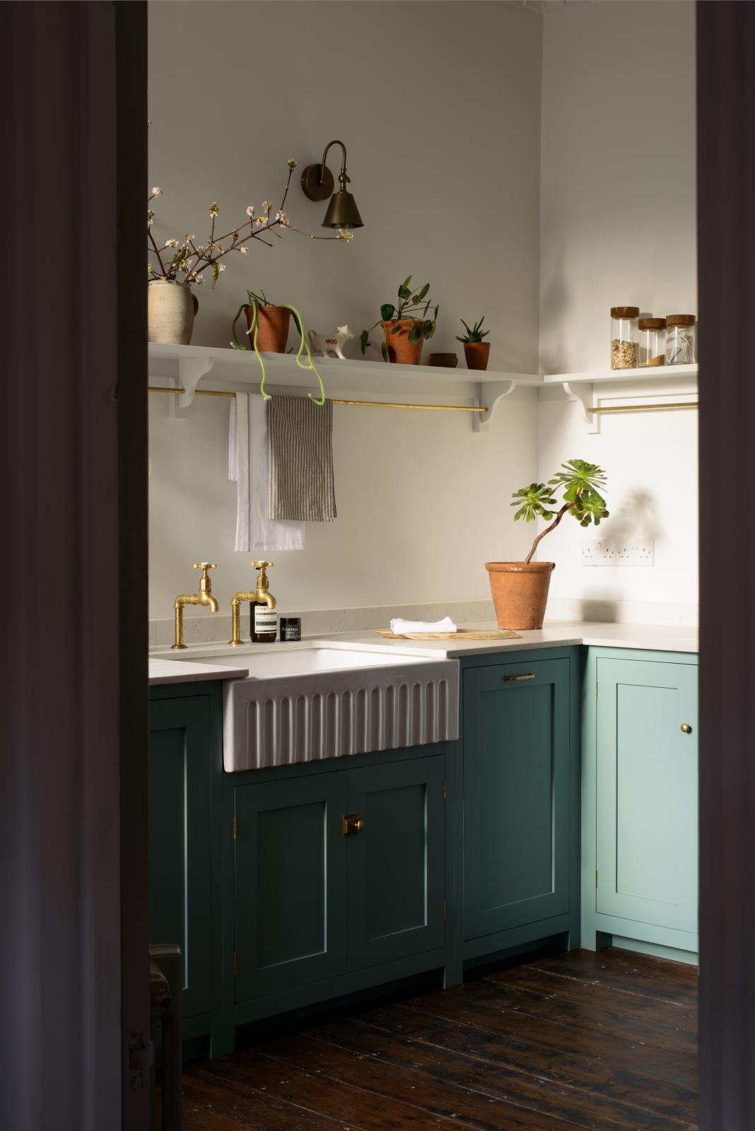 An open door in a well-preserved house reveals a small Trinity Blue scullery room, featuring a Carrara marble countertop, a single Belfast sink with aged brass Mayan taps, and warm terracotta tiles on the floor.