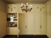 A simple corridor houses three sets of double cupboards; the left hand side is open to reveal a pantry cupboard with a microwave. Naturally decorated with a tea towel, artisan ceramics, bread, eggs, potatoes, and an antique chandelier, creating a clean and organised storage space.