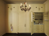 A simple corridor houses three sets of double cupboards; the right hand side is open to reveal a wine pantry cupboard with spice and bottle racks. Naturally decorated with a tea towel, wine glasses, a collection of artisan wine, and an antique chandelier, creating a clean and organised storage space.