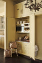 An oblique view of a simple corridor that houses three sets of double cupboards; the left-hand side is open, revealing a pantry cupboard with a microwave. Naturally decorated with a tea towel, artisan ceramics, bread, eggs, potatoes, and an antique chandelier, creating a clean and organised storage space.