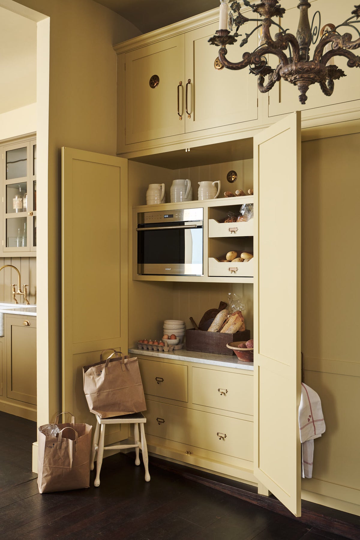 An oblique view of a simple corridor that houses three sets of double cupboards; the left-hand side is open, revealing a pantry cupboard with a microwave. Naturally decorated with a tea towel, artisan ceramics, bread, eggs, potatoes, and an antique chandelier, creating a clean and organised storage space.