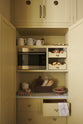A simple corridor that houses three sets of double cupboards; the left-hand side is open, revealing a pantry cupboard with a fitted microwave, a Carrara marble shelf, and a top cupboard with brass ventilation grilles. Naturally decorated with a little label holders, tea towel, artisan ceramics, bread, eggs, potatoes, and an antique chandelier, creating a clean and organised storage space.
