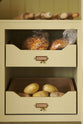 A simple corridor that houses three sets of double cupboards; the left-hand side is open, revealing a pantry cupboard with a fitted microwave, a Carrara marble shelf, and a top cupboard with brass ventilation grilles. Naturally decorated with a little label holders, tea towel, artisan ceramics, bread, eggs, potatoes, and an antique chandelier, creating a clean and organised storage space.