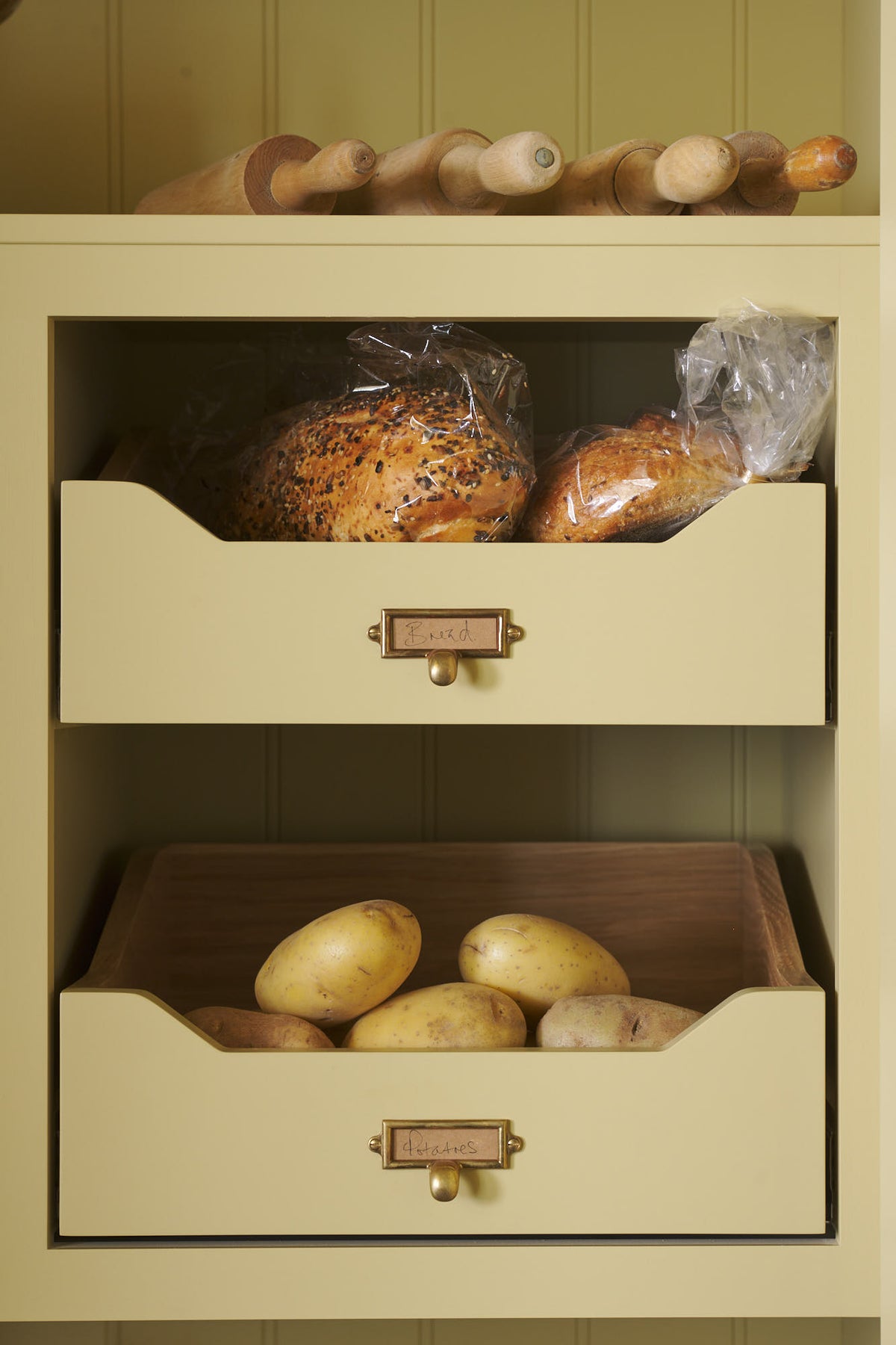 A simple corridor that houses three sets of double cupboards; the left-hand side is open, revealing a pantry cupboard with a fitted microwave, a Carrara marble shelf, and a top cupboard with brass ventilation grilles. Naturally decorated with a little label holders, tea towel, artisan ceramics, bread, eggs, potatoes, and an antique chandelier, creating a clean and organised storage space.