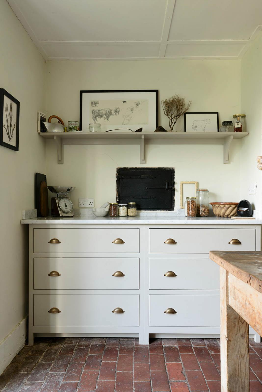 A quiet corner of a small and quaint mushroom-coloured Shaker kitchen features a sink run with Carrara marble worktop. Beautifully decorated with vintage furniture such as a large solid oak table, a silver scale, and assorted bric-a-brac, it creates a lovely social space full of character.