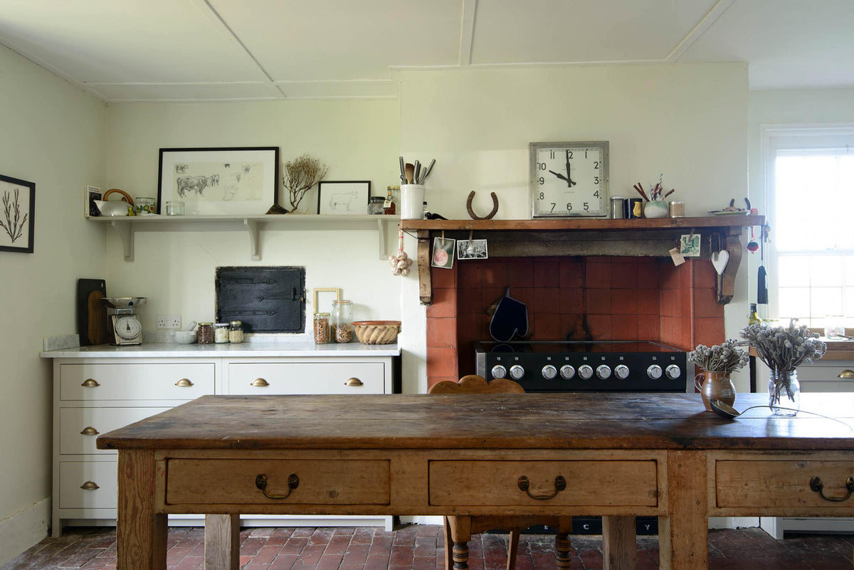 A small and quaint mushroom-coloured Shaker kitchen features a sink run made up of a mix of stave hardwood and Carrara marble worktops, a shelf, and a black stove nestled inside the original building’s chimney breast. Beautifully decorated with vintage furniture such as a large solid oak table, a silver scale, and assorted bric-a-brac, it creates a lovely social space full of character.