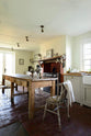 An oblique view of a small and quaint mushroom-coloured Shaker kitchen features a sink run made up of a mix of stave hardwood and Carrara marble worktops, a shelf, and a black stove nestled inside the original building’s chimney breast. Beautifully decorated with vintage furniture such as a large solid oak table, a silver scale, and assorted bric-a-brac, it creates a lovely social space full of character.