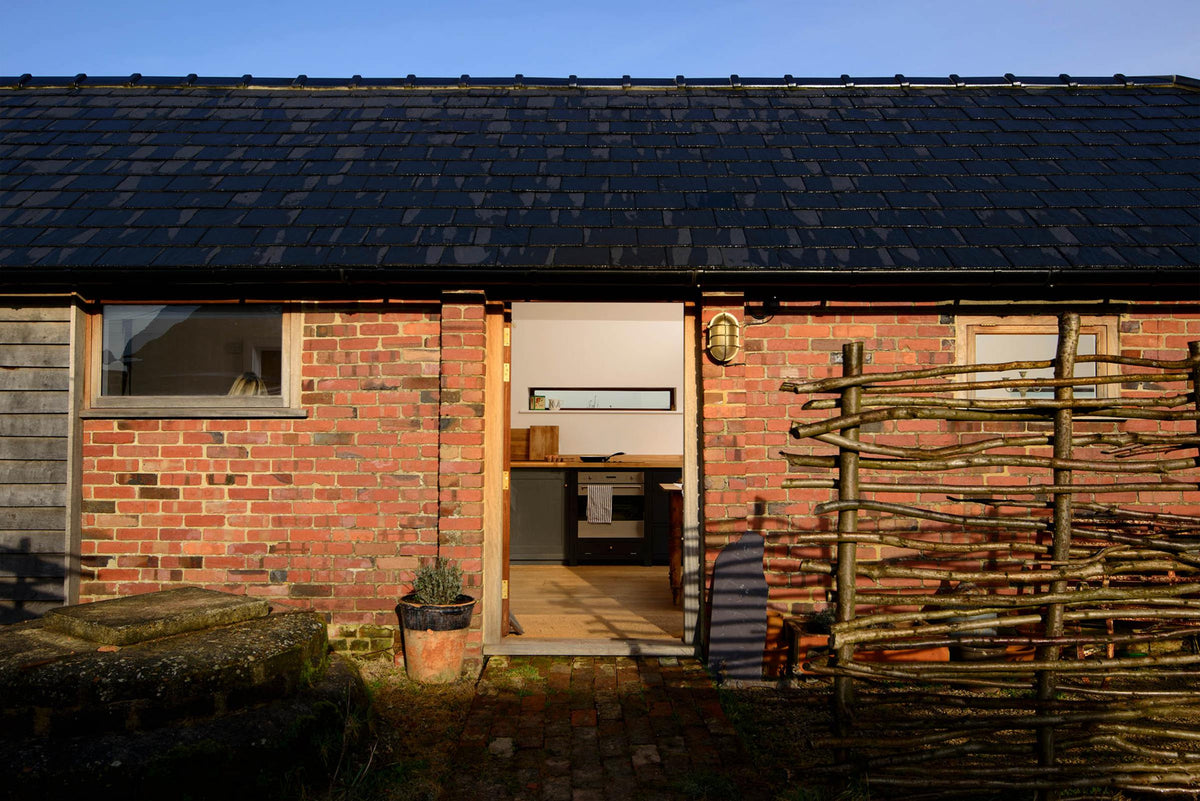 A lovely bungalow barn features a renovated roof, raw red bricks, and a handmade wooden fence. Through the open front door is a small and humble pantry-blue Shaker kitchen.