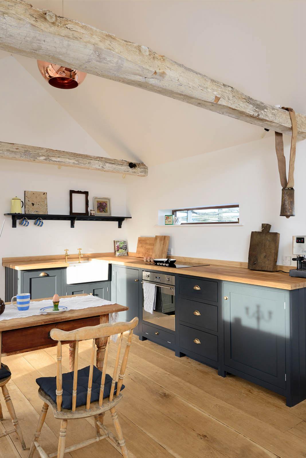 An oblique view of a small and humble pantry-blue Shaker kitchen features an L-shaped sink and cooker run with a stave hardwood worktop, a single Belfast sink, and aged brass Mayan taps. Beautifully decorated with a livestock bell attached on the roof’s wooden beams, vintage furniture, framed still-life paintings, and assorted bric-a-brac, creates a heartwarming family farm kitchen space.
