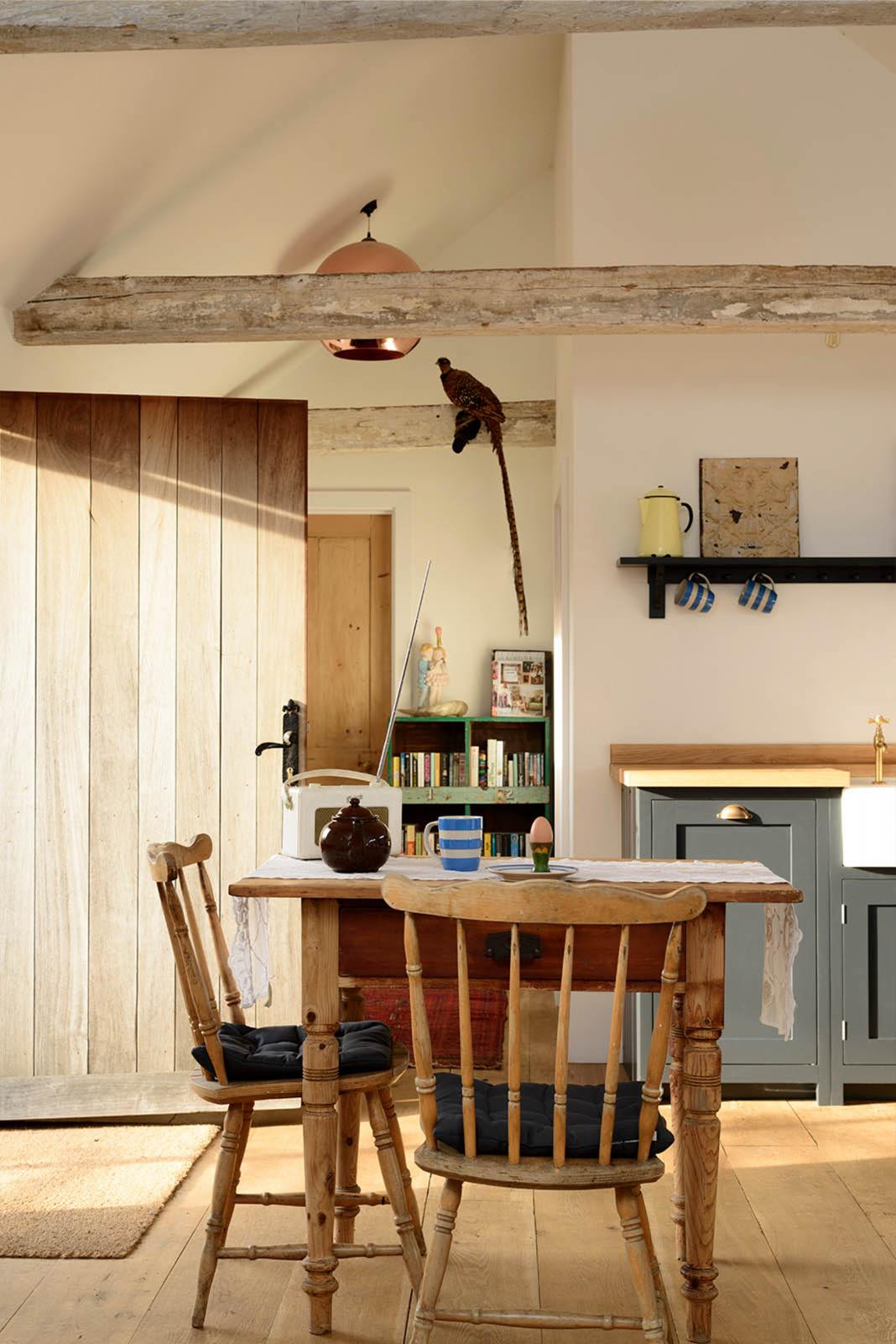 A small and humble pantry-blue Shaker kitchen features an L-shaped sink and cooker run with a stave hardwood worktop, a single Belfast sink, and aged brass Mayan taps. Beautifully decorated with a taxidermy bird sitting on the roof’s wooden beams, vintage furniture, books, and assorted bric-a-brac, creates a heartwarming family farm kitchen space.