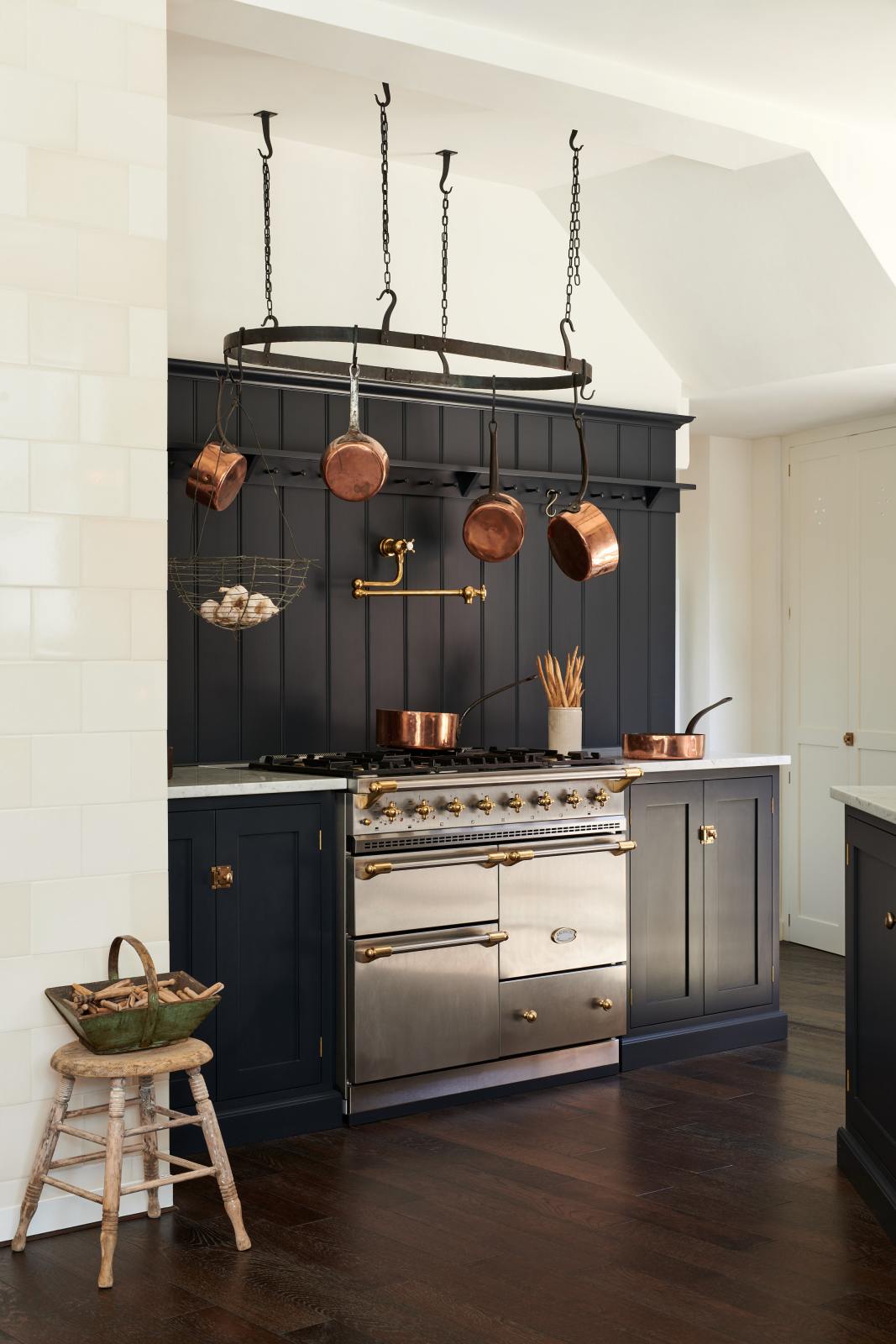 A large pantry blue shaker kitchen features a sink and cooker run with Carrara marble worktop, a silver gas cooker, and a shelf with pegs. Beautifully decorated with vintage furniture, copper pans, a pot hanger, and an aged brass pot filler tap, the kitchen presents a thoughtfully curated mix of collected pieces that fit together effortlessly.