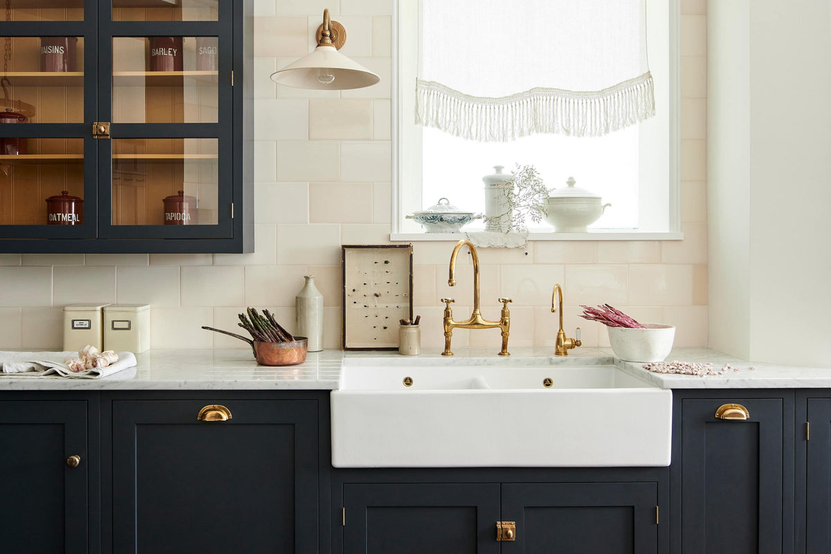A large pantry blue shaker kitchen features a sink and cooker run with Carrara marble worktop, a double Belfast sink, an aged brass Ionian tap, wall cabinets with Scullery yellow interior, and cream subway splashback tiles. Beautifully decorated with vintage items, artisan ceramics, an antique bug display, and a deVOL Heirloom task light, the kitchen presents a thoughtfully curated mix of collected pieces that fit together effortlessly.