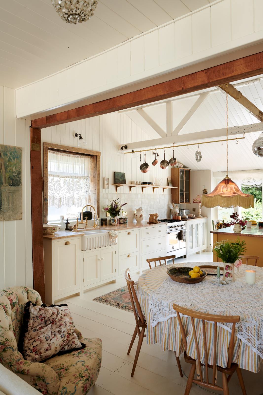 A beach house kitchen with Shaker cupboards, wooden floors and panelled walls, all painted in a fresh and bright off-white. There is a window behind the tactile ribbed sink and a brass mixer tap with a pretty patina. A table is just peeking into shot on the right-hand side, making this a very comfortable and social family kitchen.