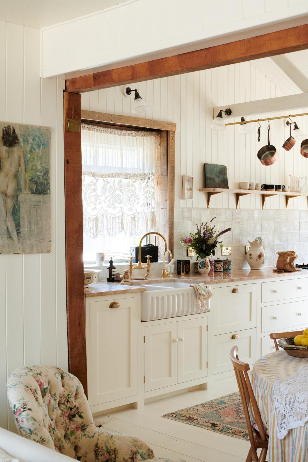 A beach house kitchen with Shaker cupboards, wooden floors and panelled walls, all painted in a fresh and bright off-white. There is a window behind the tactile ribbed sink and a brass mixer tap with a pretty patina. A table is just peeking into shot on the right-hand side, making this a very comfortable and social family kitchen.