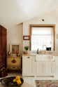 Looking straight towards the back wall of a bohemian-style kitchen that has tongue and groove panelled walls, solid floorboards, and a run of simple Shaker furniture, all painted in a creamy white shade. A mustardy yellow prep table sits in the centre of the room, creating a galley-style working area. The kitchen is fitted with soft pink marble worktops and styled with a collection of vintage pots and pans.