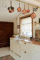 A diagonal view of a sink run in a Bohemian-inspired beach house kitchen, featuring white cabinets with luminescent marble worktops and a fluted sink with two brass taps. An oriental-style carpet covers the floor, and a dark red patterned curtain partially covers one side of the wall. Bronze pots in various sizes hang from the brass rail on the ceiling, enhancing a more vintage mood.