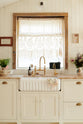 A sink runs along a Bohemian-inspired beach house kitchen, featuring white cabinets with luminescent marble worktops and a fluted sink with two brass taps. Behind the sink, sunlight pours through the window with s a wood frame. A short sheer ruffled curtain with a floral print on the window adds an extra touch of charm to the kitchen.