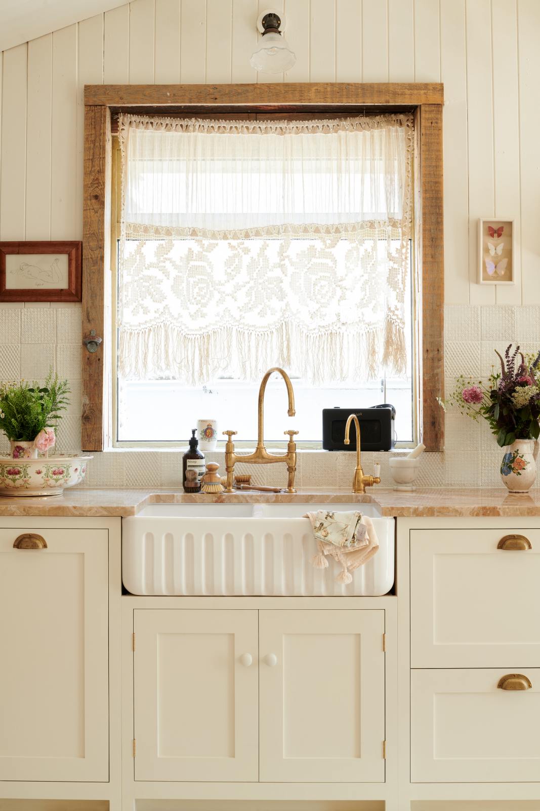 A sink runs along a Bohemian-inspired beach house kitchen, featuring white cabinets with luminescent marble worktops and a fluted sink with two brass taps. Behind the sink, sunlight pours through the window with s a wood frame. A short sheer ruffled curtain with a floral print on the window adds an extra touch of charm to the kitchen.