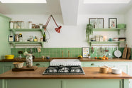 A vibrant pea-green Shaker kitchen features a sink run and a kitchen island with a gas cooker, both topped with a reclaimed iroko school countertop, matching green square splashback tiles, shelves with brass hanging rails, and a double oven cabinet with drawers. Beautifully decorated with artisan ceramics, dried ingredients in a glass jar, oranges, and a clamped red lamp, creates a friendly and welcoming kitchen.