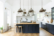 A tall pantry blue shaker kitchen features a sink and cooker run with a Silestone worktop, complemented by a large kitchen island topped with solid oak and glazed cupboards. The cooker run sits in an alcove with white splashback tiles, topped by a shaker shelf displaying assorted ceramic and wooden bric-a-bracs. Rusty metal hanging lights in the centre add an industrial touch to the space.