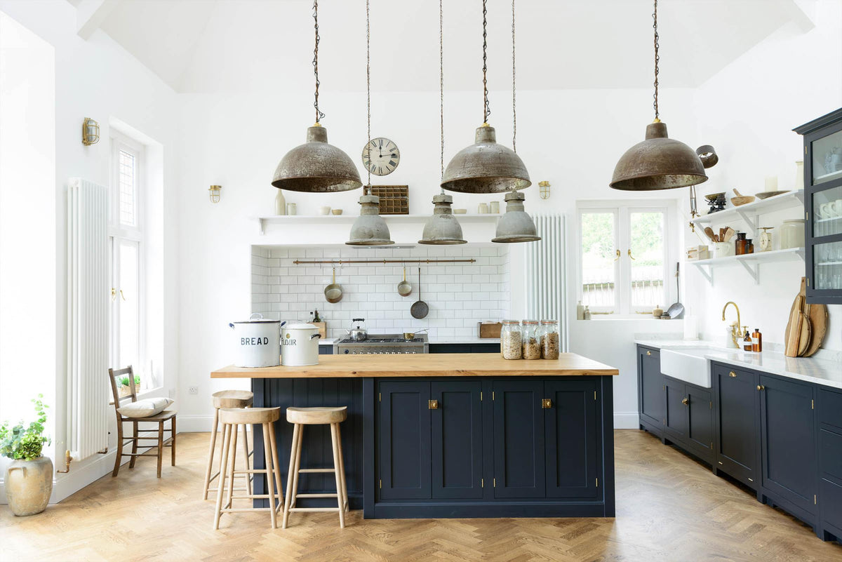 A tall pantry blue shaker kitchen features a sink and cooker run with a Silestone worktop, complemented by a large kitchen island topped with solid oak and glazed cupboards. The cooker run sits in an alcove with white splashback tiles, topped by a shaker shelf displaying assorted ceramic and wooden bric-a-bracs. Rusty metal hanging lights in the centre add an industrial touch to the space.