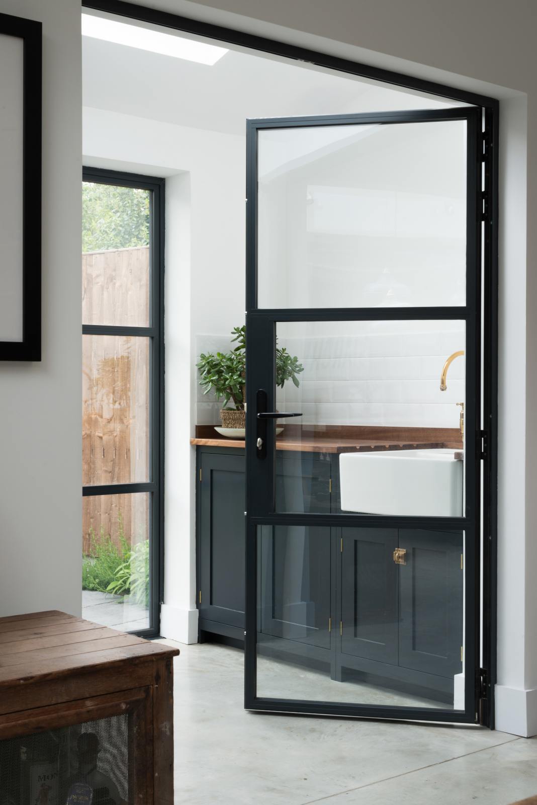 Past the black-framed French doors is a small sink run with a single sink, an aged brass Ionian tap, and a solid oak worktop. Adorned with a luscious green house plant, it creates a warm and social space.