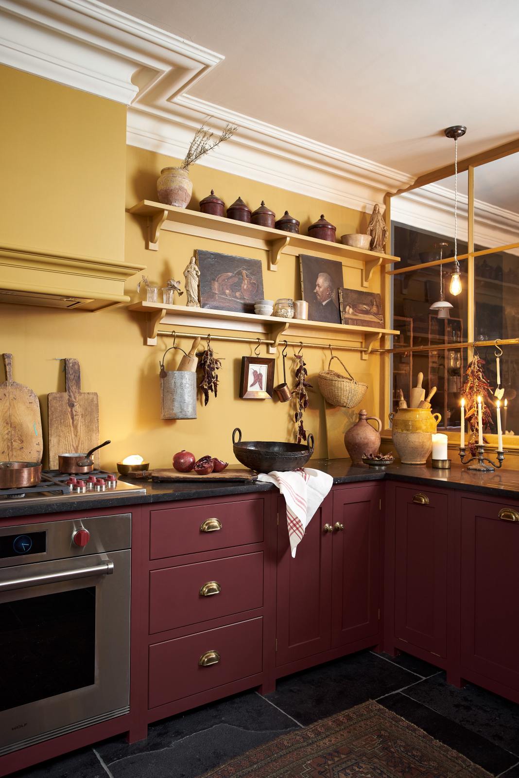 The corner of a busy and vibrant Refectory Red Shaker kitchen features an L-shaped sink and cooker run with a Belgian blue limestone worktop, shelves with brass rails, and a classic Shaker canopy. Adorned with vintage items, beloved ceramics, still-life paintings, wooden chopping boards, and candles creates a bright and wonderfully social kitchen space.