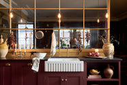 A busy and vibrant Refectory Red Shaker kitchen features an L-shaped sink and cooker run with a Belgian blue limestone worktop, a single fluted Belfast sink, and aged brass Mayan taps. Adorned with vintage items, beloved ceramics, still-life paintings, dried chillies, red lettuce, and an arrangement of berry branches, creates a bright and wonderfully social kitchen space.