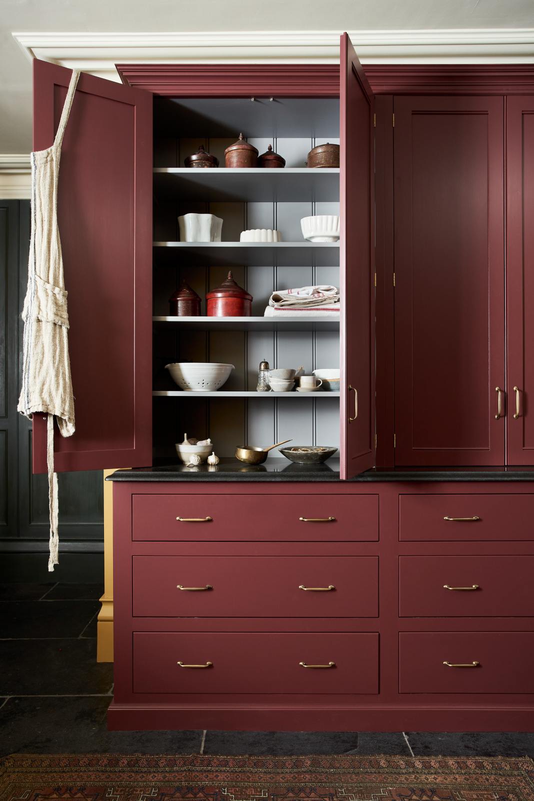 A busy and vibrant Refectory Red Shaker kitchen features a glazed larder with drawers and a Belgian blue limestone worktop. Adorned with vintage items, beloved ceramics, tea towels and bulbs of garlic, creates a bright and wonderfully social kitchen space.