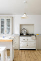 A minimalist, mushroom-coloured shaker kitchen includes a cooker around a chimney breast, a white gas stove, and a porcelain splashback, complemented by a freestanding table and a metal chair near the window.