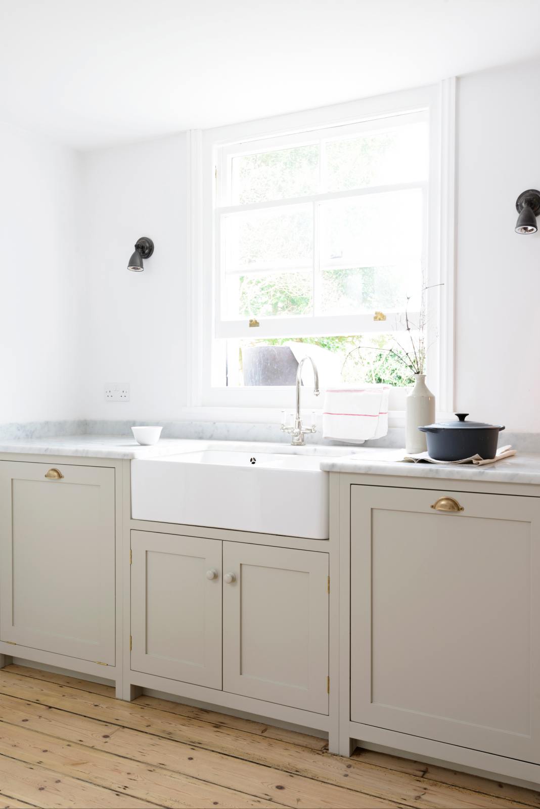 An angled view of a simple mushroom-coloured shaker kitchen features a marble worktop, a double Belfast sink with an antique silver Ionian tap, and glazed cabinets.