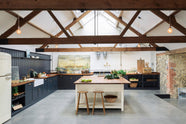 A large, renovated cattle shed has been transformed into a spacious pantry-blue Shaker kitchen, featuring an L-shaped cooker and sink run, along with a large white linen kitchen island topped with a stave hardwood worktop. Beautifully decorated with aged brass taps, Helen stools, luscious green vegetables, vintaged artisan ceramics, lilac tulips and abstract paintings.