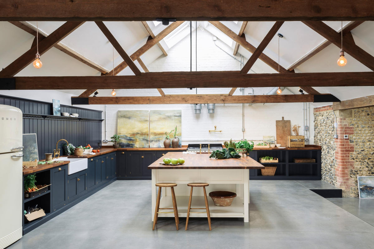 A large, renovated cattle shed has been transformed into a spacious pantry-blue Shaker kitchen, featuring an L-shaped cooker and sink run, along with a large white linen kitchen island topped with a stave hardwood worktop. Beautifully decorated with aged brass taps, Helen stools, luscious green vegetables, vintaged artisan ceramics, lilac tulips and abstract paintings.