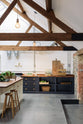 Nestled between an L-shaped cooker and sink run and a large white linen kitchen island topped with a stave hardwood worktop, the spacious pantry-blue Shaker kitchen has transformed a large, renovated cattle shed. Beautifully decorated with an aged brass pot filler tap, Helen stools, luscious green vegetables, vintaged artisan ceramics, dried flowers and an abstract painting.