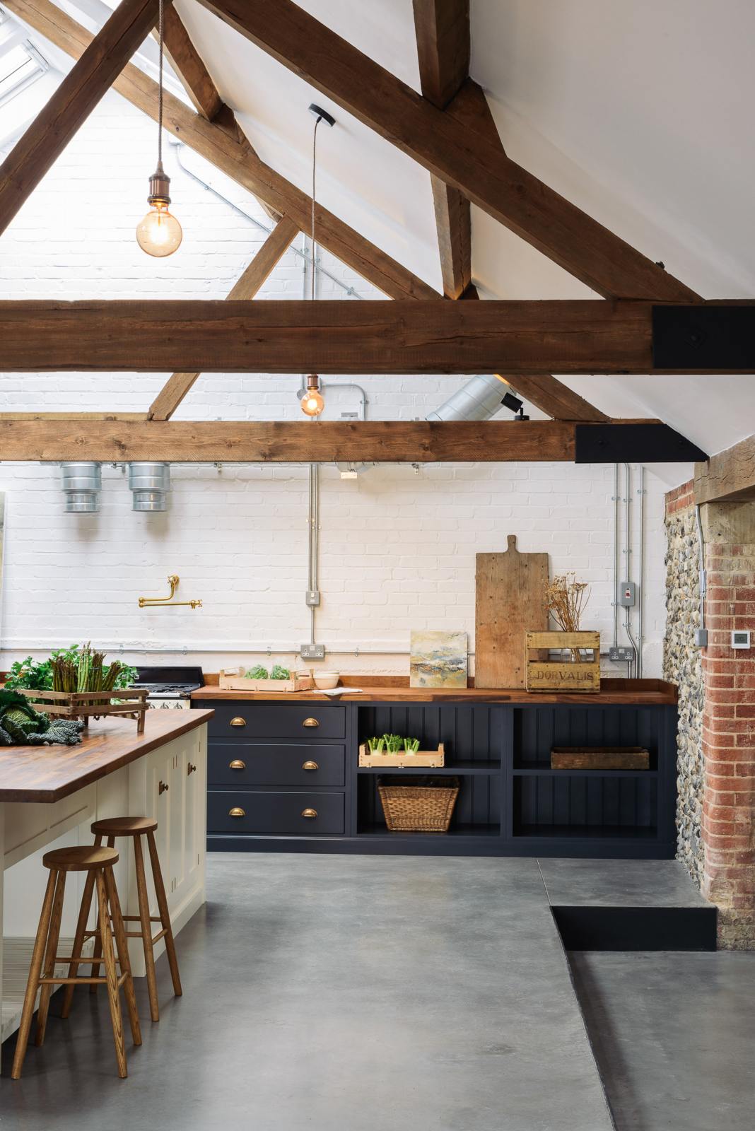 Nestled between an L-shaped cooker and sink run and a large white linen kitchen island topped with a stave hardwood worktop, the spacious pantry-blue Shaker kitchen has transformed a large, renovated cattle shed. Beautifully decorated with an aged brass pot filler tap, Helen stools, luscious green vegetables, vintaged artisan ceramics, dried flowers and an abstract painting.