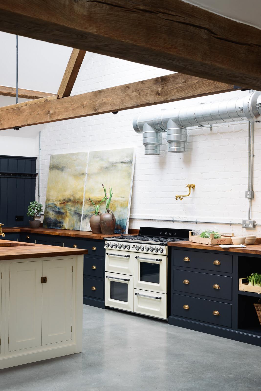 An oblique view of a large, renovated cattle shed has been transformed into a spacious pantry-blue Shaker kitchen, featuring an L-shaped cooker and sink run, along with a large white linen kitchen island topped with a stave hardwood worktop. Beautifully decorated with an aged brass pot filler tap, a white gas stove, luscious green vegetables, vintage artisan ceramics, lilac tulips and abstract paintings.